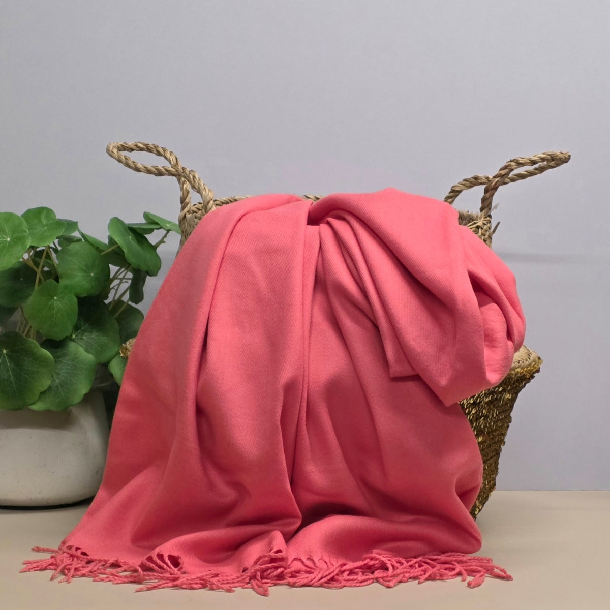 A pink fabric with fringed edges draped over a woven straw basket, with a potted green plant on the left against a light grey background.