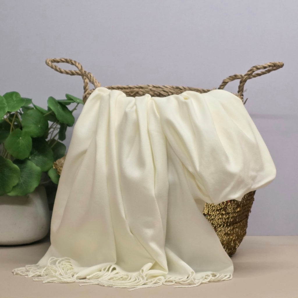 A cream fabric with fringed edges draped over a woven straw basket, with a potted green plant on the left against a light grey background.