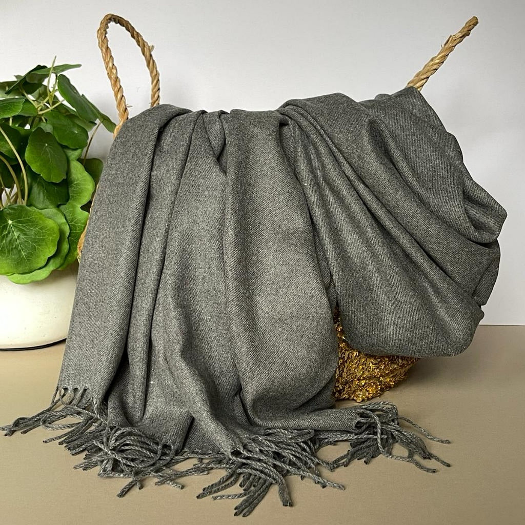 Gray throw blanket with fringe draped over a woven basket on a neutral background