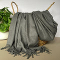 Gray throw blanket with fringe draped over a woven basket on a neutral background
