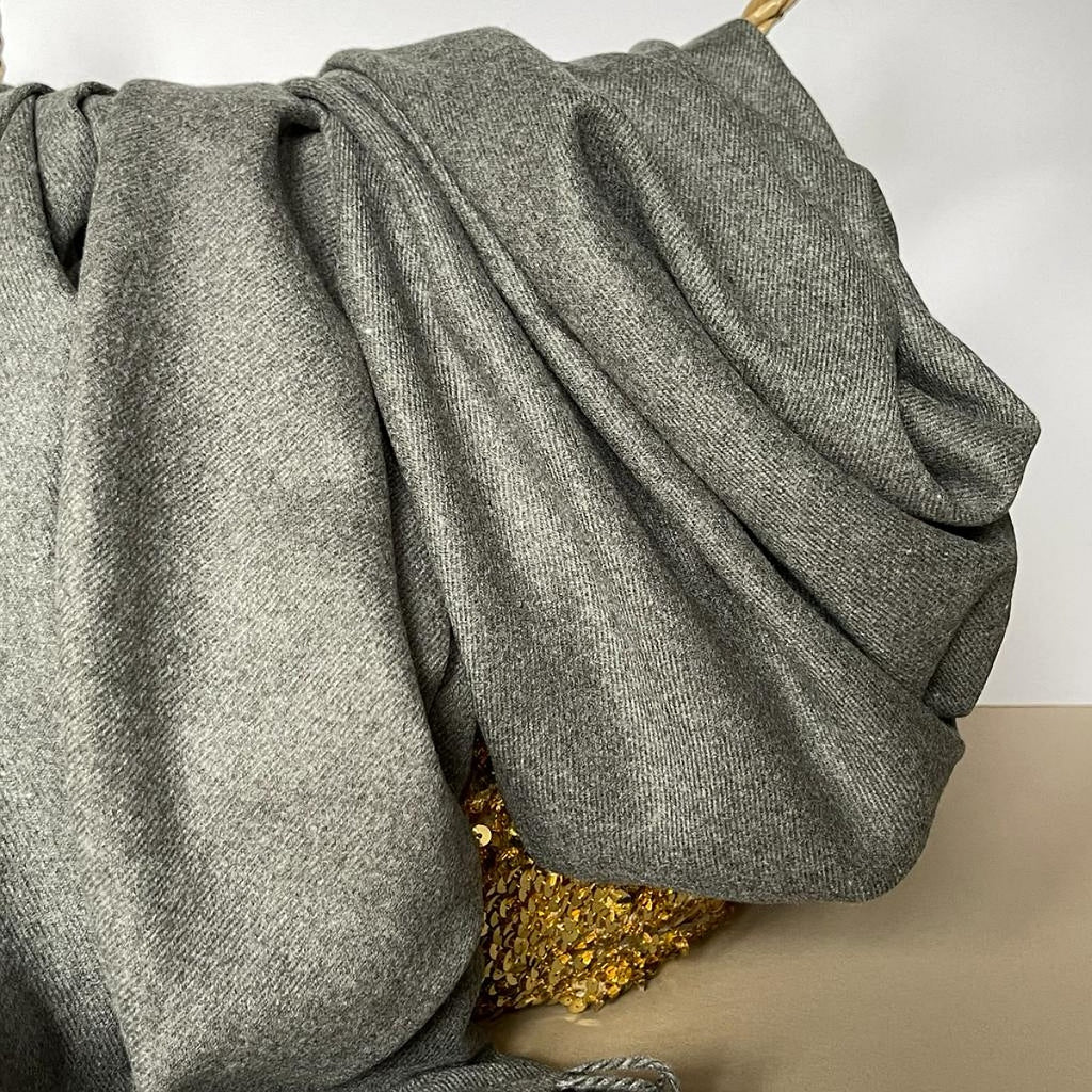 A close-up of a draped gray woven fabric, likely a scarf or throw, with a textured herringbone pattern, resting on a gold sequined surface against a light background.