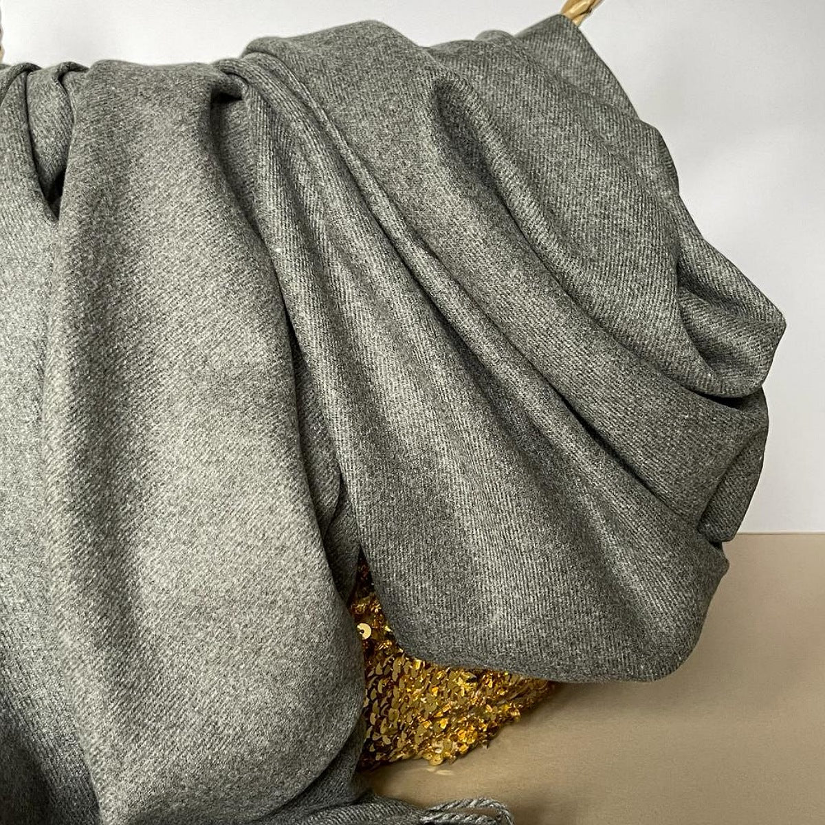 A close-up of a draped gray woven fabric, likely a scarf or throw, with a textured herringbone pattern, resting on a gold sequined surface against a light background.