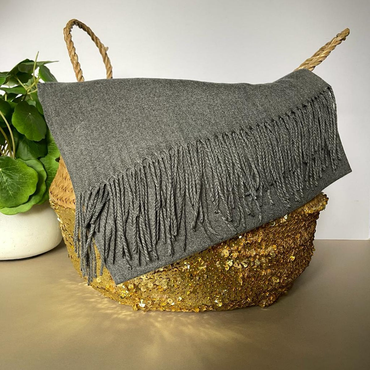 A gray knit throw blanket with long fringe, draped over a woven basket with rope handles, set on a tabletop with a potted plant in the background.