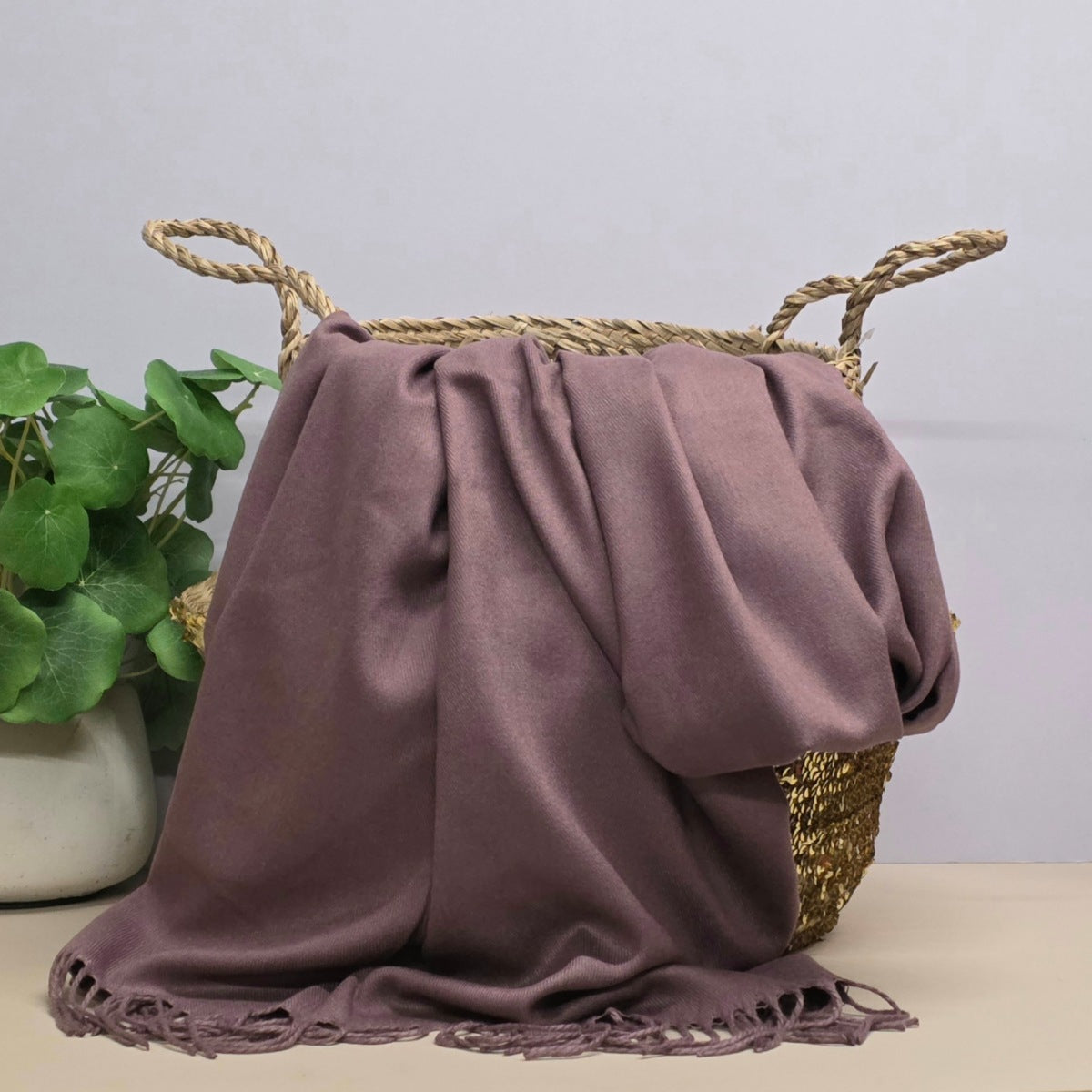 A dusty mauve purple shawl with fringed edges draped over a woven straw basket with rope handles, set next to a green potted plant on a light beige surface against a light grey background.