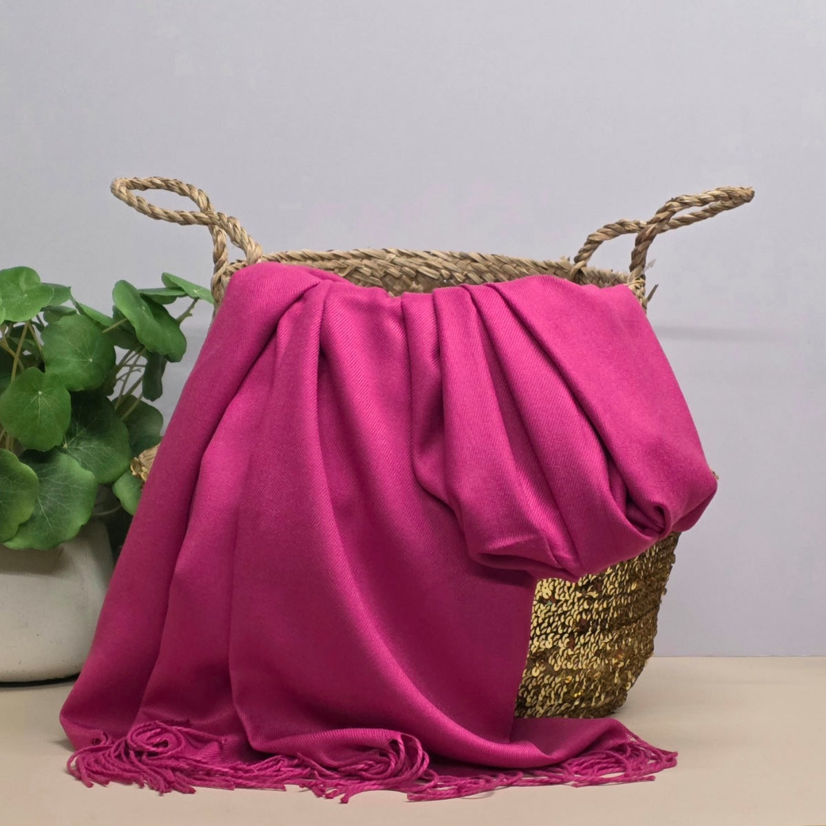 A dark pink fabric with fringed edges draped over a woven straw basket, with a potted green plant on the left against a light grey background.
