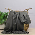 A grey fabric with fringed edges draped over a woven straw basket, with a potted green plant on the left against a light grey background.
