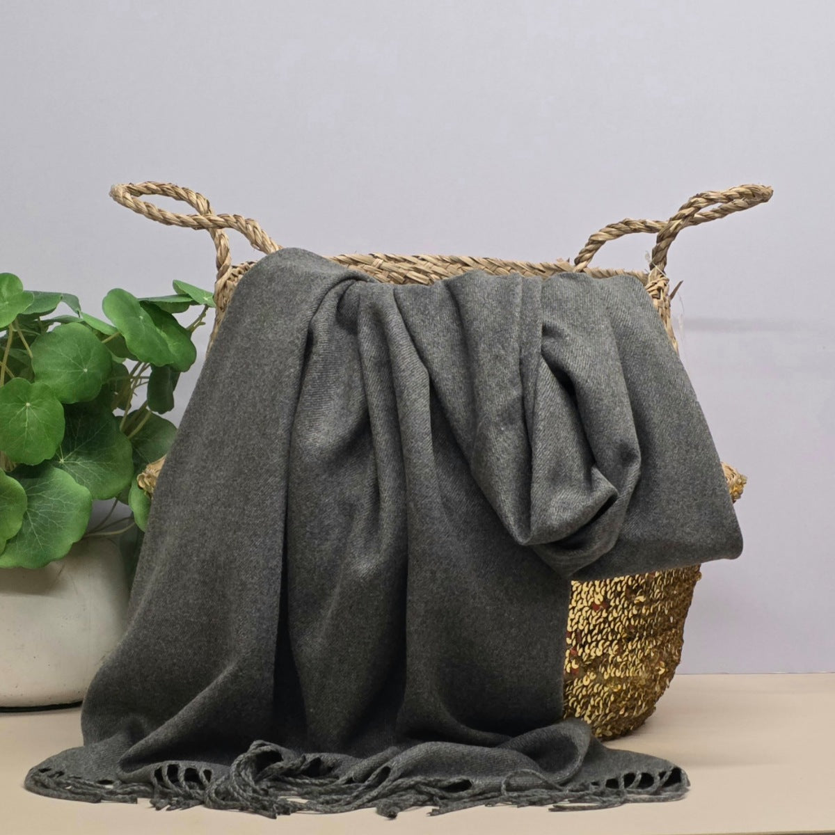 A grey fabric with fringed edges draped over a woven straw basket, with a potted green plant on the left against a light grey background.