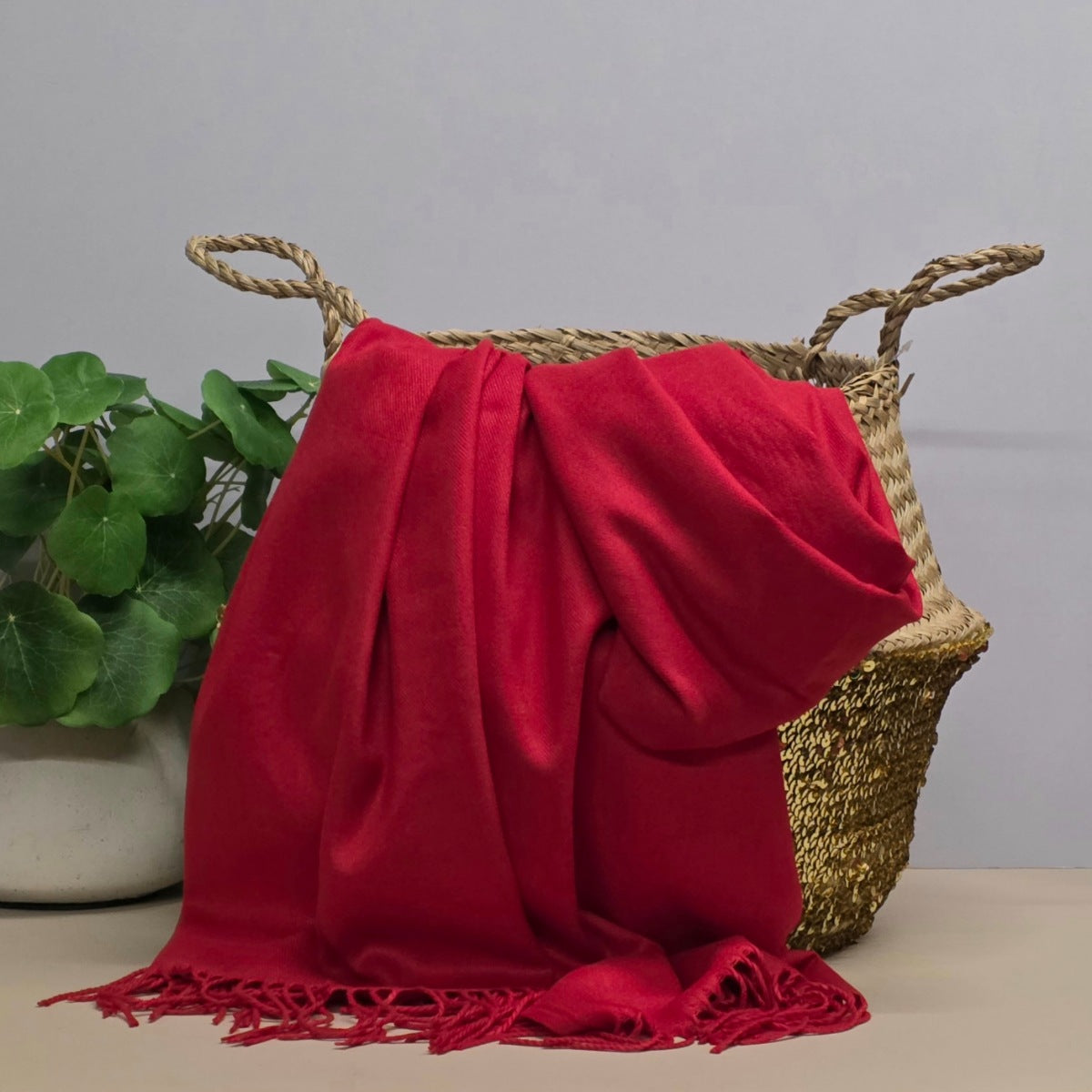 A red fabric with fringed edges draped over a woven straw basket, with a potted green plant on the left against a light grey background.