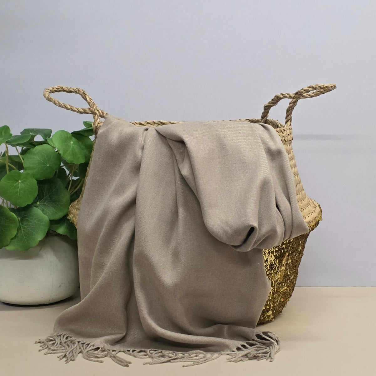 A draped taupe-beige knit throw blanket with fringe, styled over a woven straw tote bag with a gold sequin bottom, next to a potted green plant on a light beige surface against a pale grey background.