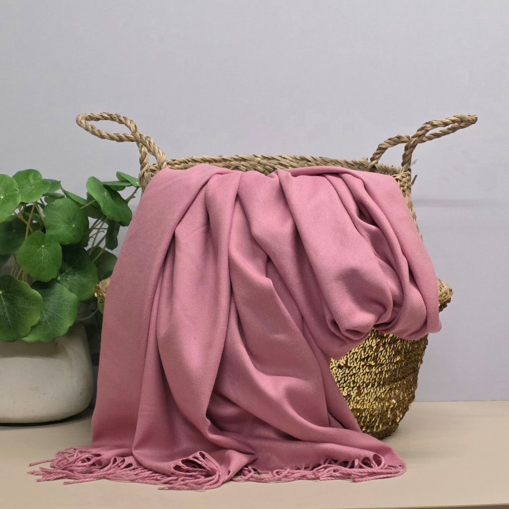 A dusty pink throw blanket with fringed edges draped over a woven basket, set on a light beige surface next to a green potted plant against a light grey wall.