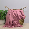 A dusty pink throw blanket with fringed edges draped over a woven basket, set on a light beige surface next to a green potted plant against a light grey wall.