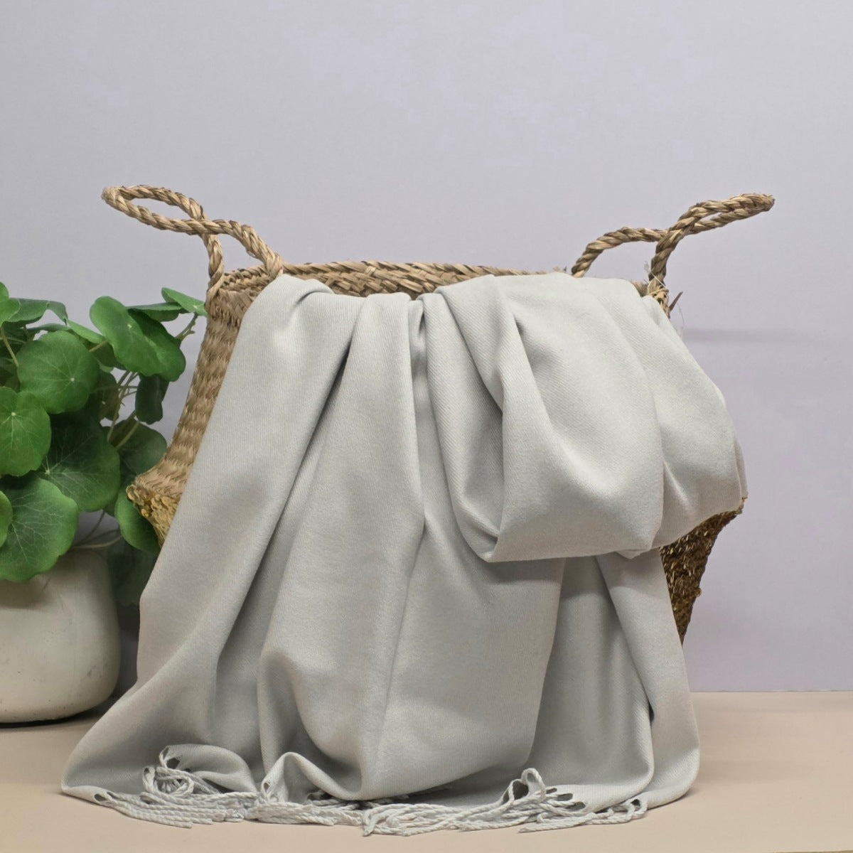 A light grey throw blanket with fringed edges draped over a woven straw basket, with a potted green plant in the background against a pale grey wall.