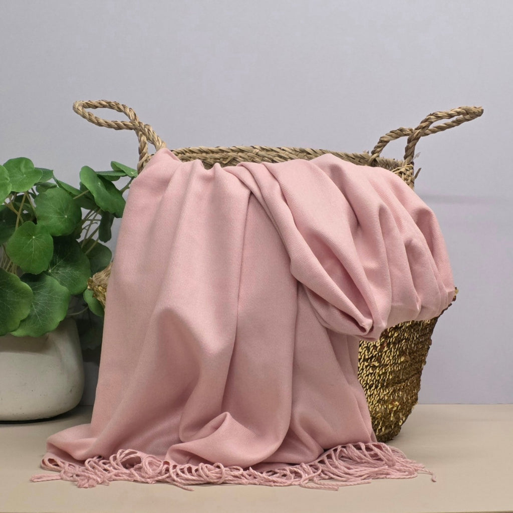 A light pink fabric with fringed edges draped over a woven straw basket, with a potted green plant on the left against a light grey background.