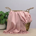 A light pink fabric with fringed edges draped over a woven straw basket, with a potted green plant on the left against a light grey background.
