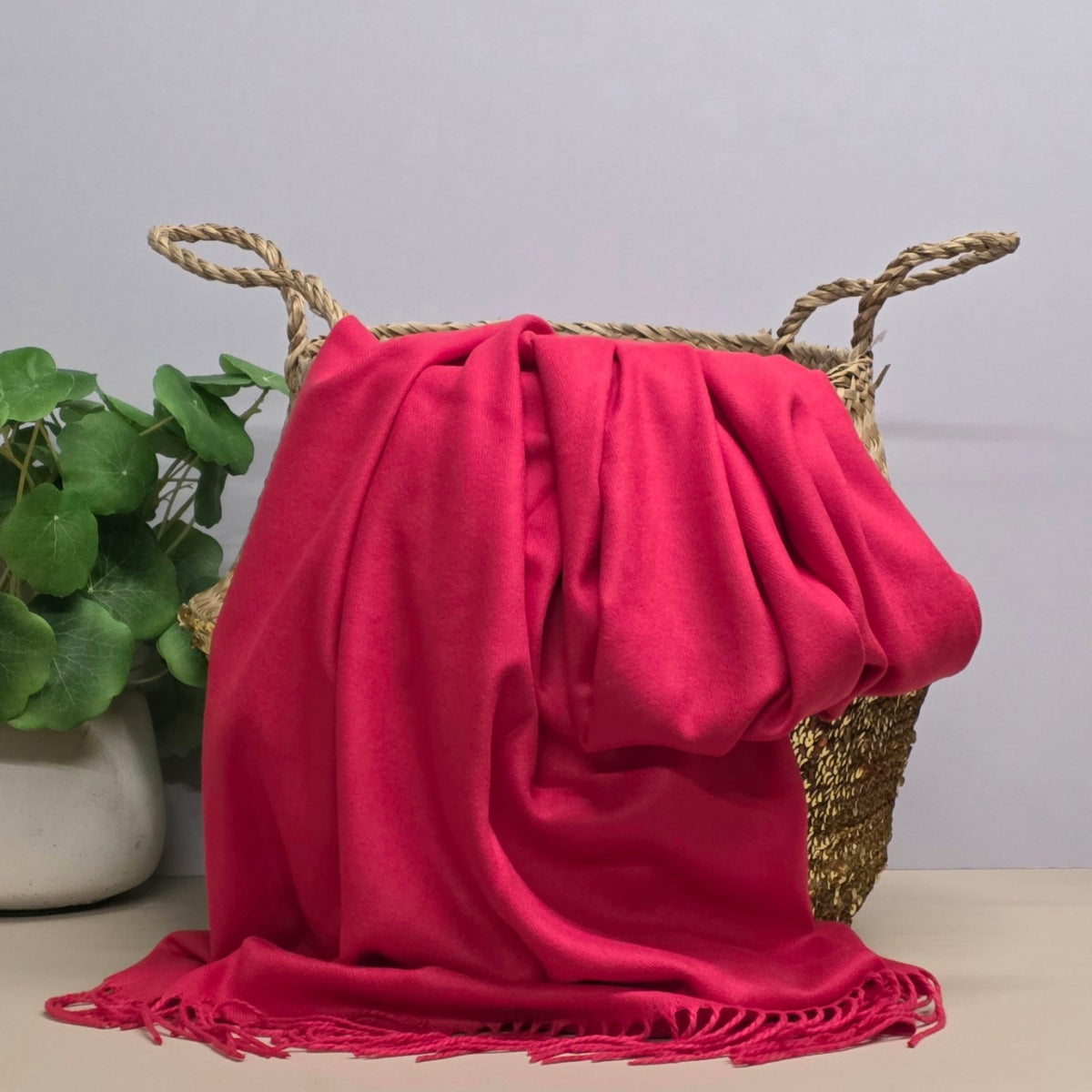 A red fabric with fringed edges draped over a woven straw basket, with a potted green plant on the left against a light grey background.