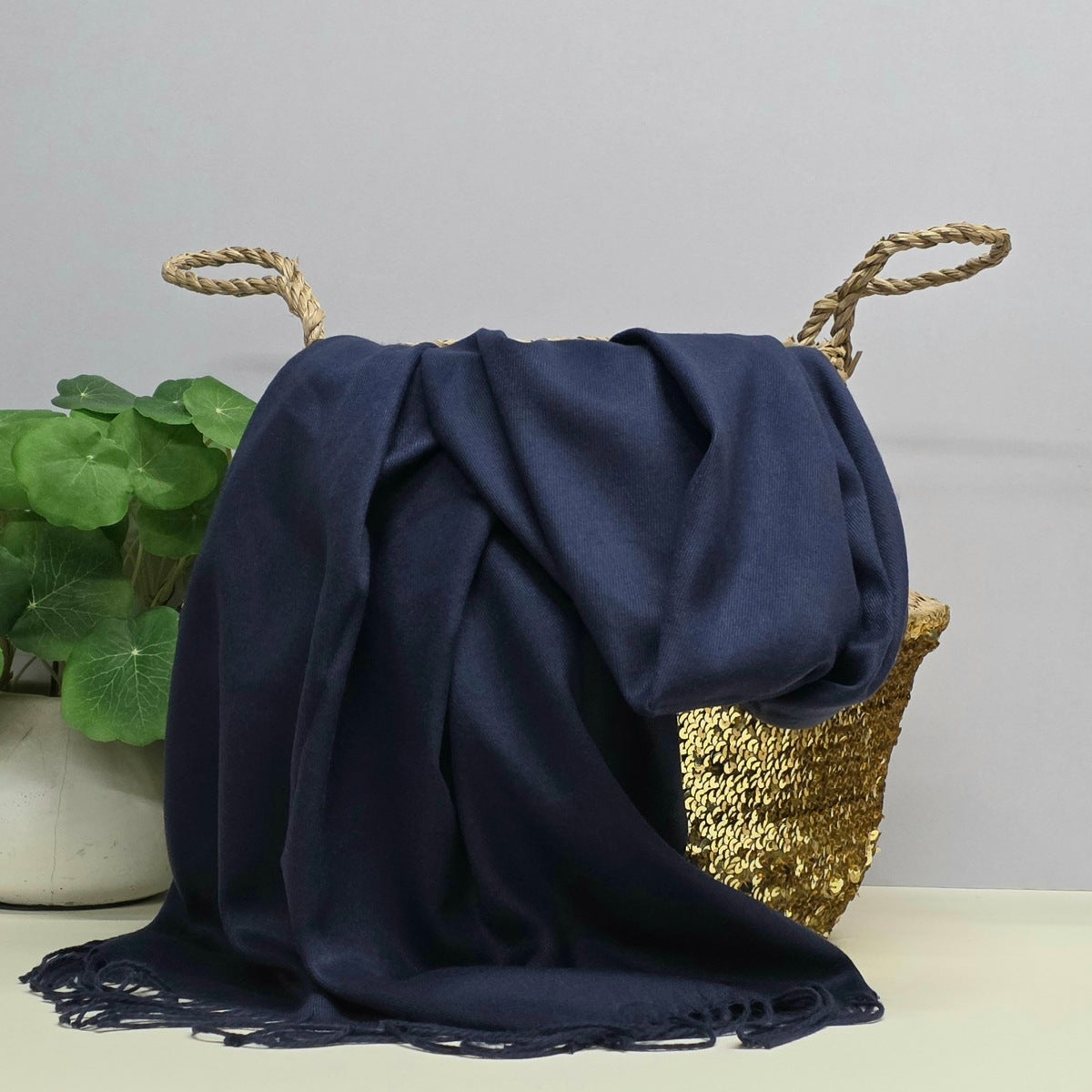 A navy blue fringed throw blanket draped over a woven straw basket with rope handles, set against a light gray background with a green potted plant on the left.