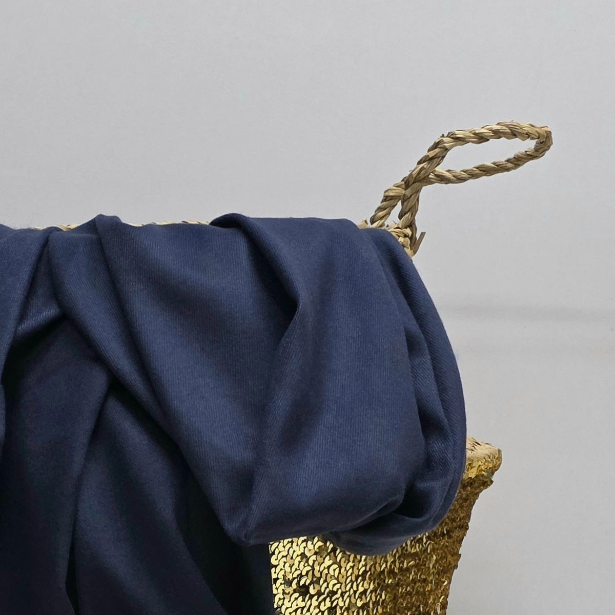 A draped navy blue fabric, likely a throw blanket or scarf, styled over a woven straw basket with a rope handle against a neutral gray background.