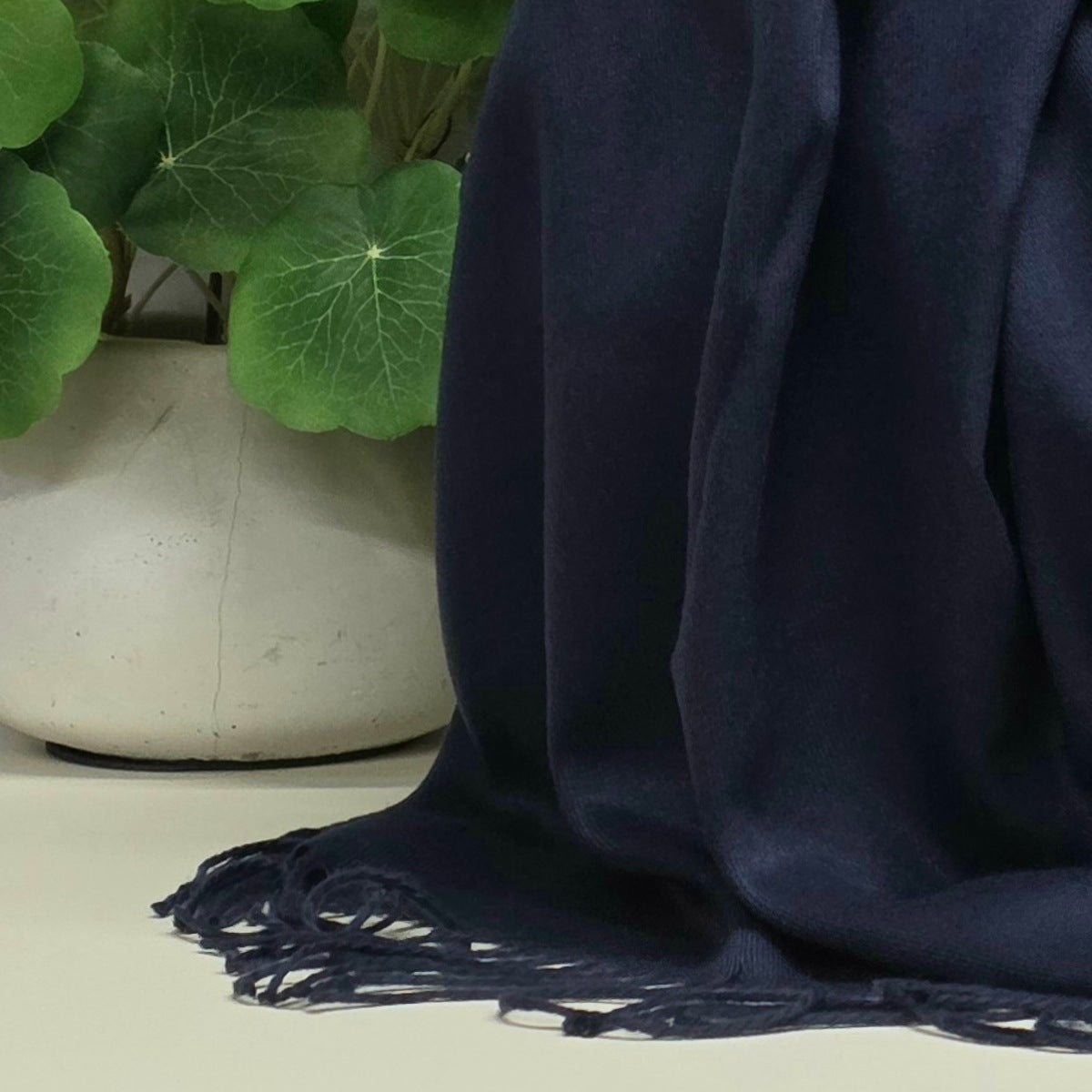 A dark navy blue pashmina shawl with fringed edges draped over a light-colored surface next to a white ceramic planter with green leaves.