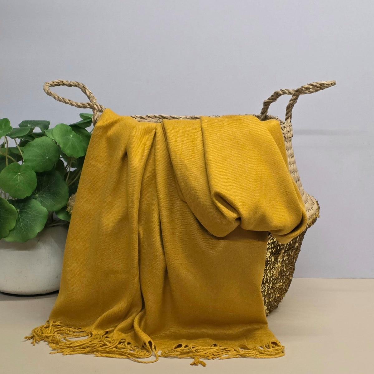 A yellow fabric with fringed edges draped over a woven straw basket, with a potted green plant on the left against a light grey background.