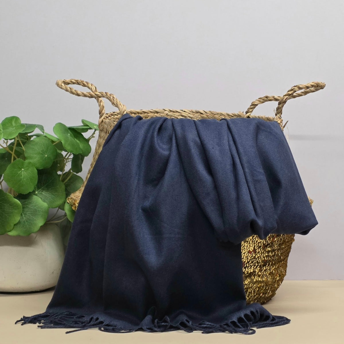 A navy throw blanket with fringed edges draped over a woven straw basket, with a potted green plant on the left against a light grey background.