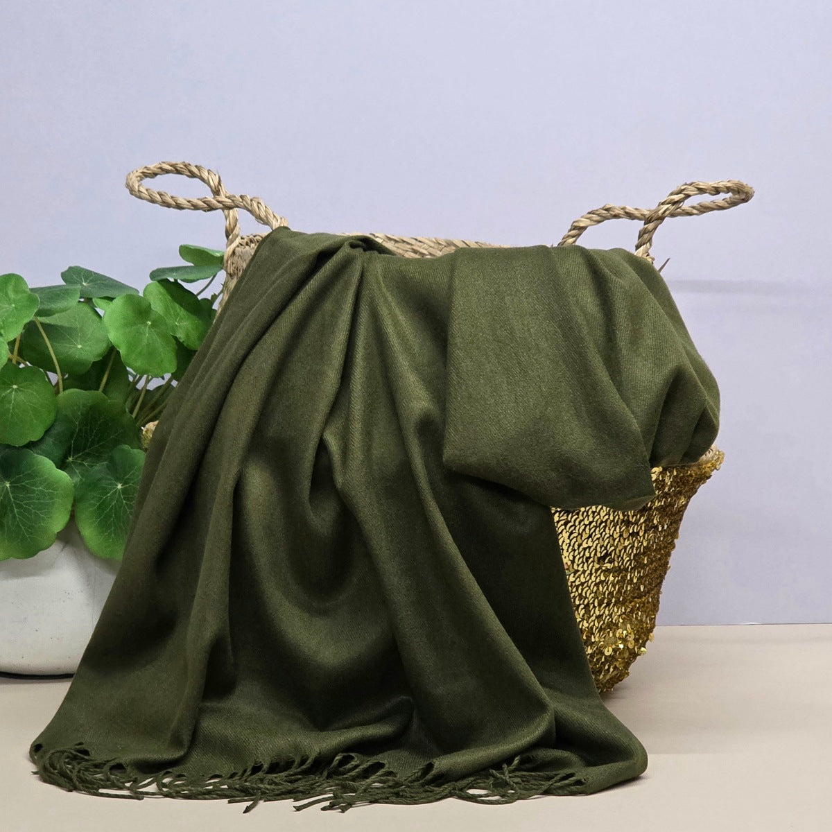 An olive green shawl with fringed edges draped over a woven straw basket, with a potted green plant on the left against a grey background.