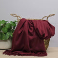 A deep burgundy throw blanket with fringed edges draped over a woven straw basket, with a potted green plant on the left against a light grey background.