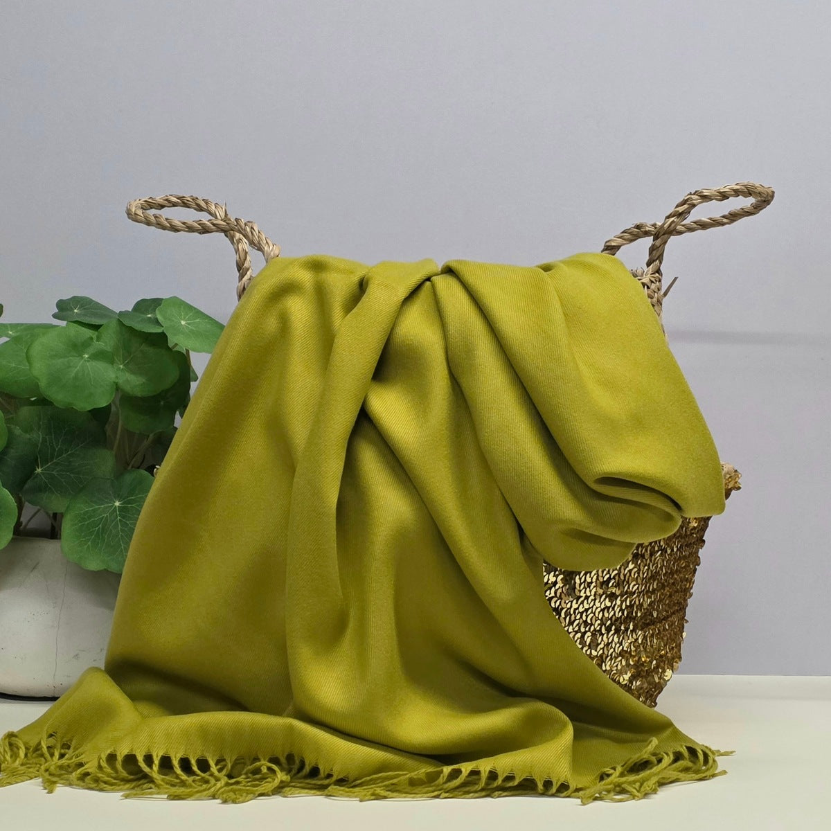 An olive green silk-like throw blanket with fringed edges draped over a woven basket with rope handles, set against a light gray background with a green plant on the left.