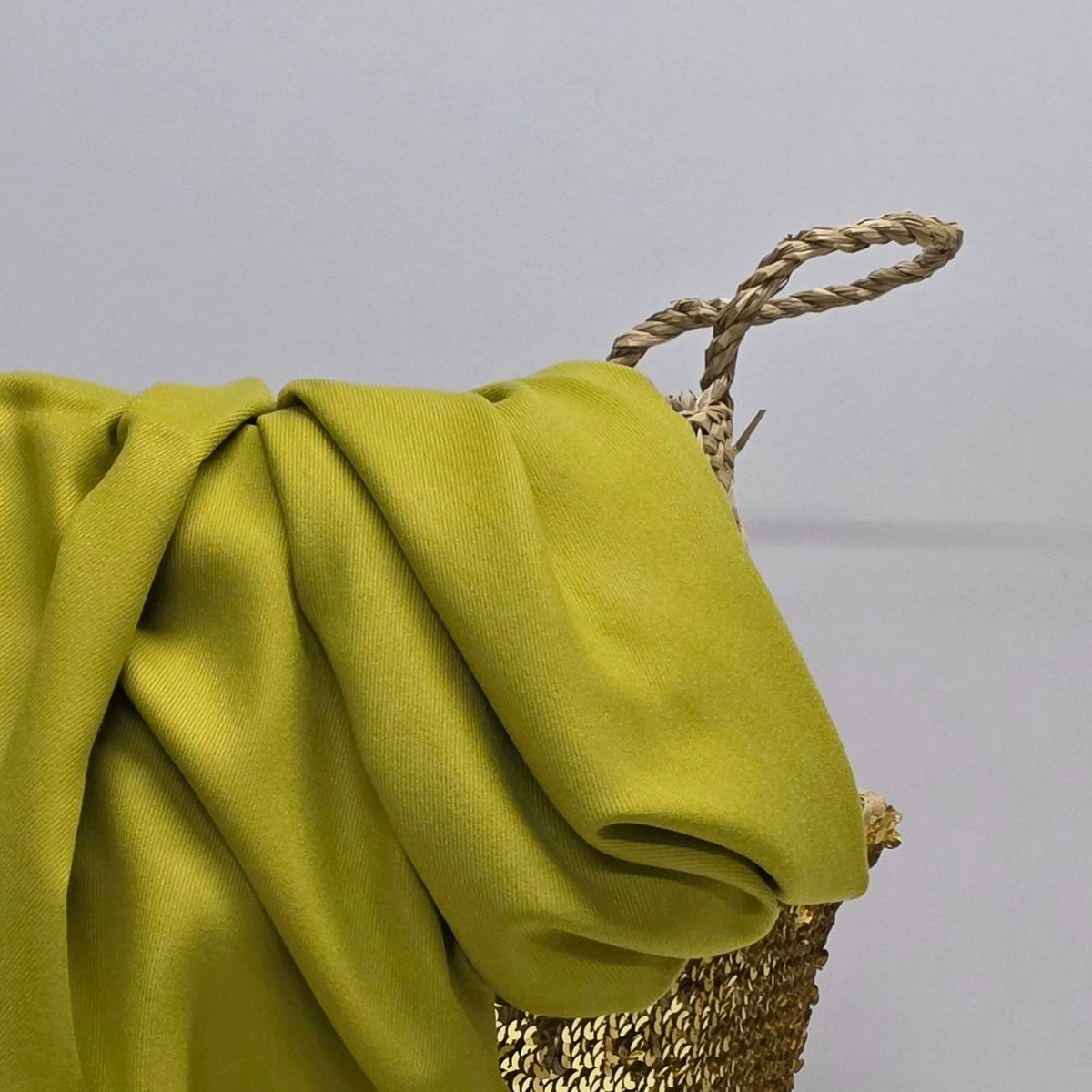 A draped olive green fabric throw blanket with a twisted rope handle, resting on a woven gold basket against a neutral gray background.