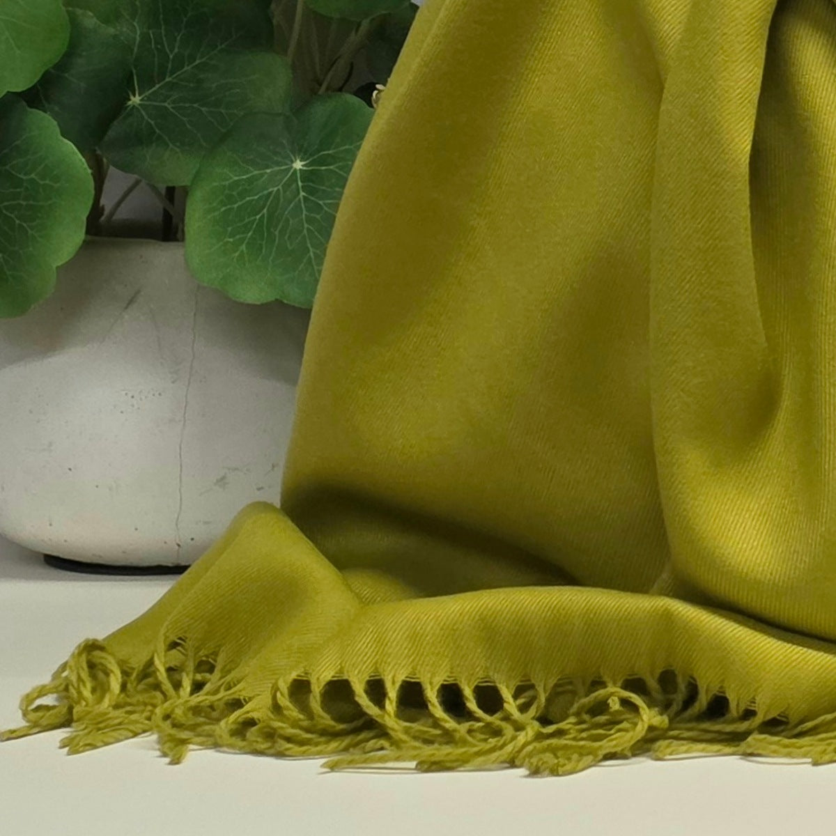 A close-up of a draped olive green scarf with fringed edges, shown next to a white ceramic pot with green leaves.
