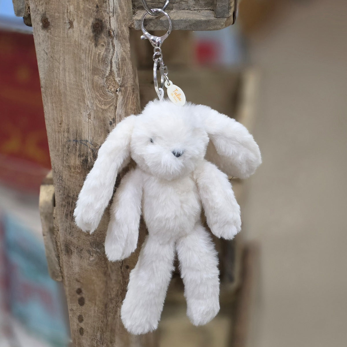 A cream plush bunny keychain hanging from a rustic wooden post. The keychain features a soft, fuzzy cream bunny with black eyes and nose, attached to a silver metal key ring and a small round tag.
