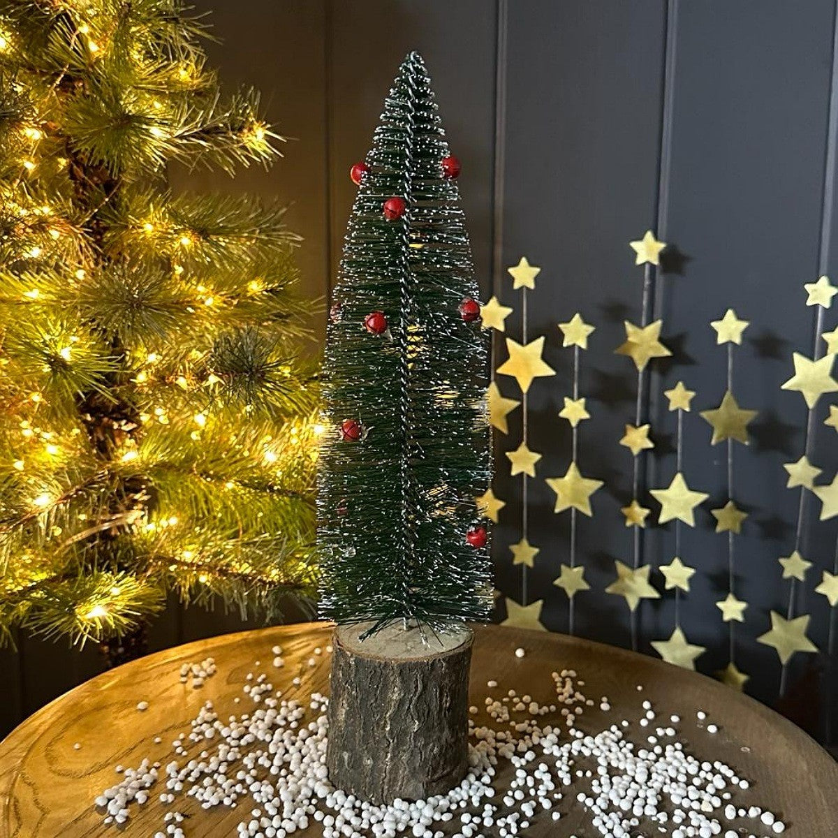 A small artificial Christmas tree with white faux snow, red bell ornaments, and a rustic wood base, displayed on a wooden surface with decorative gold stars and a warm-lit Christmas tree in the background.