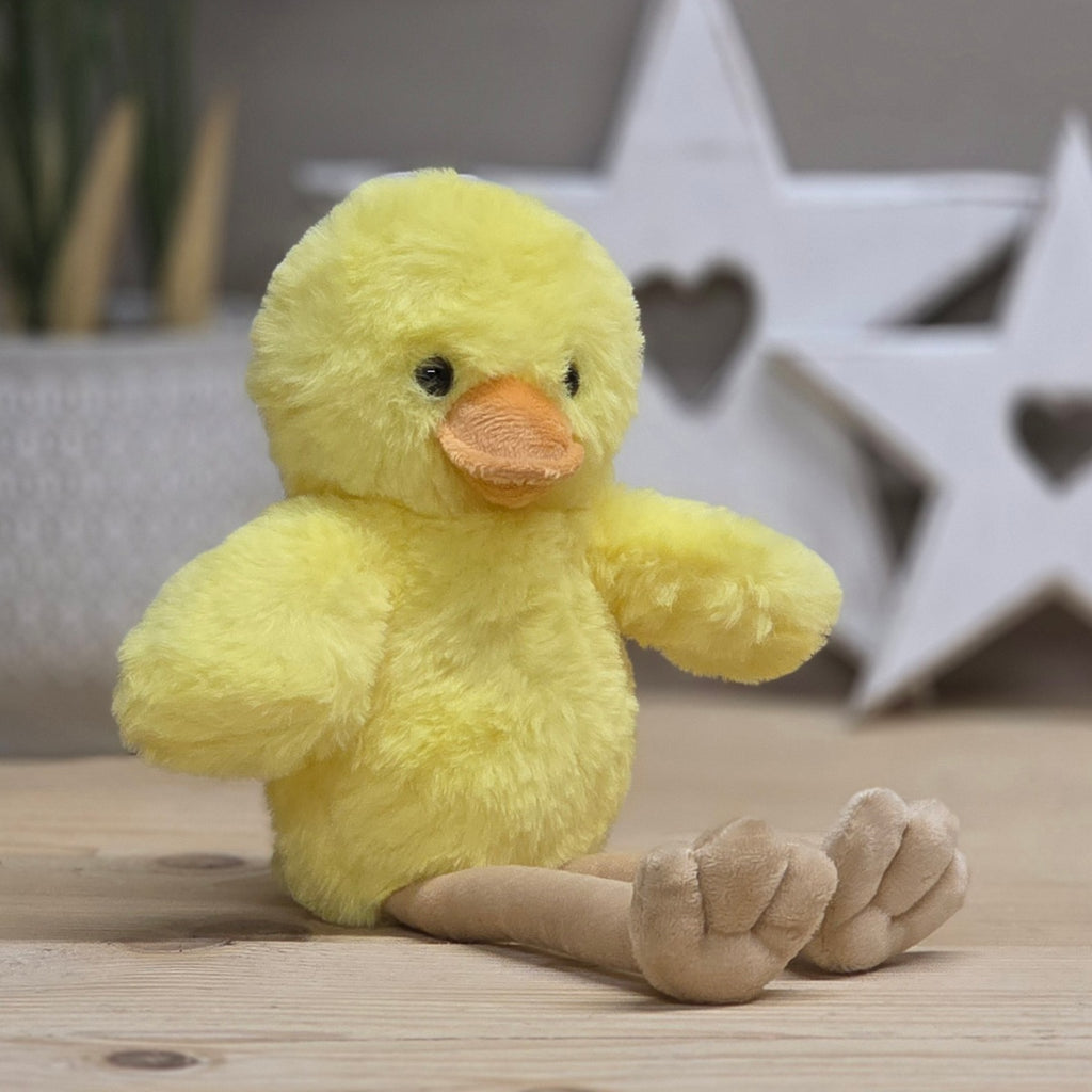 A yellow soft fuzzy plush chick with an orange beak and beige long legs, with black eyes.  On a wooden surface with a blurred star-shaped object in the background
