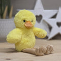 A yellow soft fuzzy plush chick with an orange beak and beige long legs, with black eyes.  On a wooden surface with a blurred star-shaped object in the background

