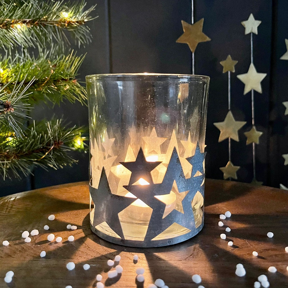 A clear glass candle holder decorated with star-shaped metal cutouts, lit from within with a warm yellow glow, set on a wooden surface with holiday decorations in the background.