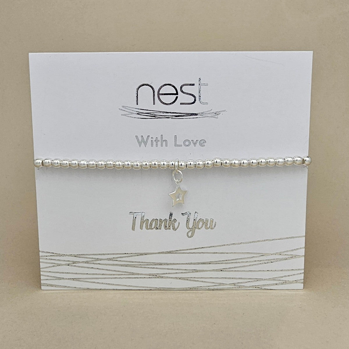 A silver beaded bracelet with a dangling star charm, presented on a white 'nest With Love Thank You' gift card against a beige background.