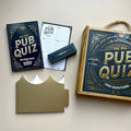 Wooden box with words 'The Big Pub Quiz' game set on a light surface with packaging and information.