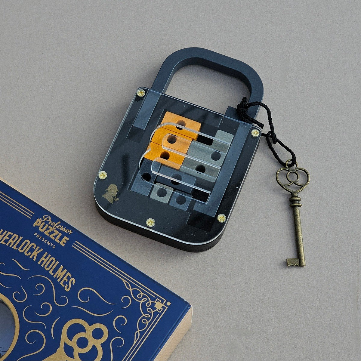 A transparent lock-shaped puzzle box with a dark handle and internal orange and grey sliding pieces, shown on a neutral grey surface next to a blue and gold Sherlock Holmes box and a small heart-shaped key.