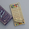 A wooden board game called Code Breaker shown with its purple box on a neutral grey background. The game board is made of light natural wood with engraved slots and text, and contains colorful game pieces in green, blue, red, yellow, and white.