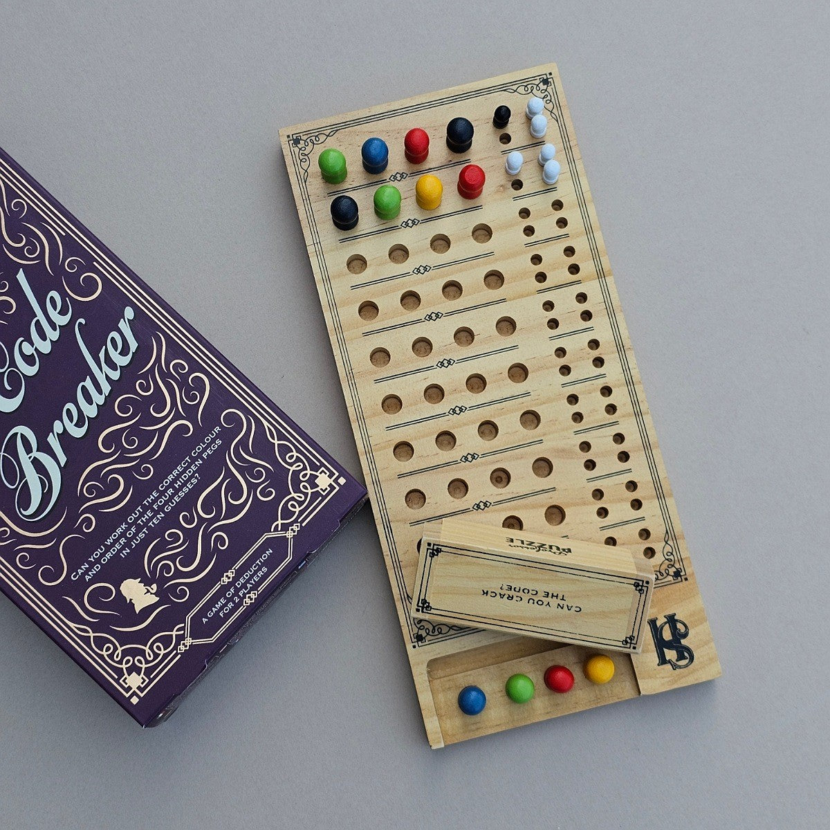 A wooden board game called Code Breaker shown with its purple box on a neutral grey background. The game board is made of light natural wood with engraved slots and text, and contains colorful game pieces in green, blue, red, yellow, and white.