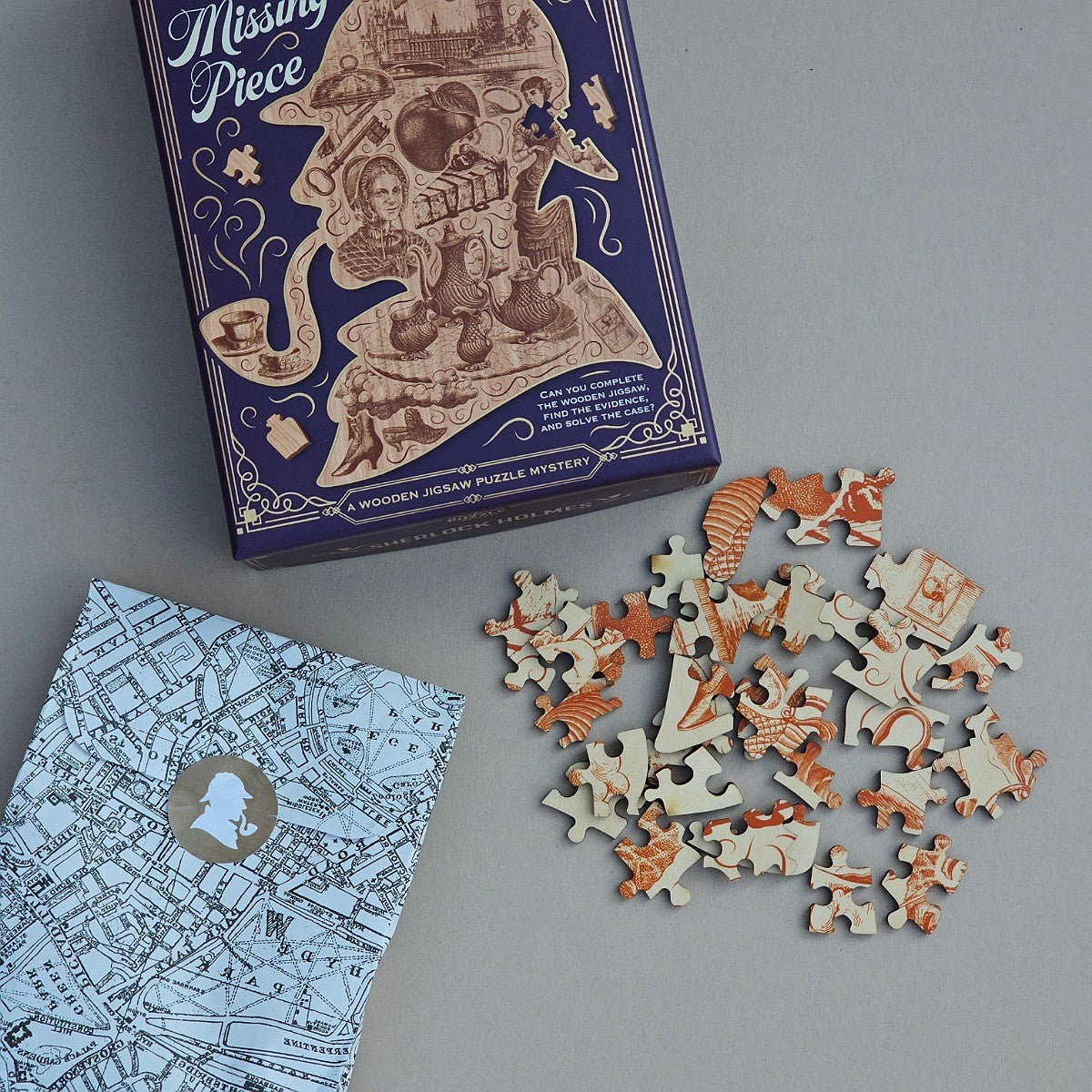 A boxed jigsaw puzzle set labeled 'Missing Piece' shown on a grey surface, with the illustrated dark blue box, a folded map-patterned booklet, and a scattered pile of cut puzzle pieces.