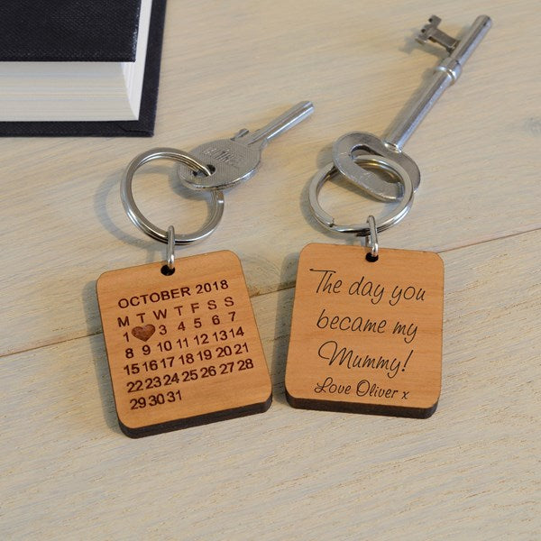 Two engraved wooden keychains with metal keys on a light wood tabletop. One keychain shows a calendar for October 2018 with a heart over the number 2, and the other says 'The day you became my Mummy! Love Oliver x'.