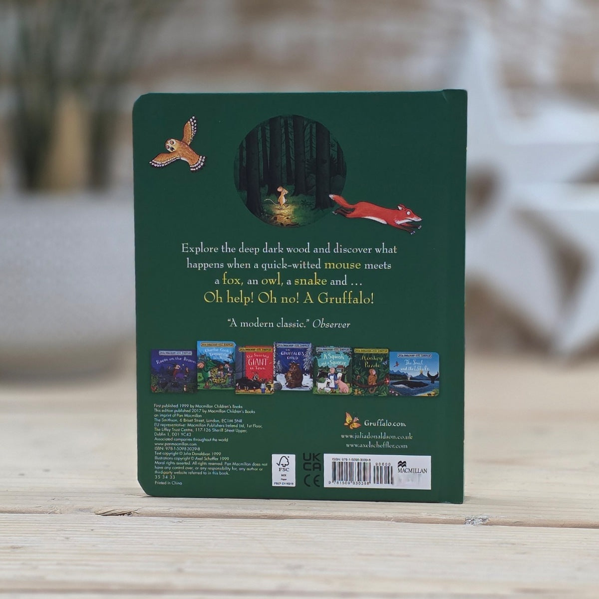 The back cover of a children's book titled 'The Gruffalo' By Julia Donaldson with a sub heading 'Oh my! Oh no! A Gruffalo'. It is standing upright on a light wooden surface with a blurred outdoor background.