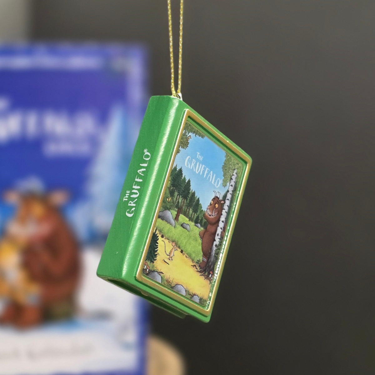 A green book-shaped ornament for The Gruffalo hanging from a gold string, with a colorful illustrated cover and a blurred book in the background.