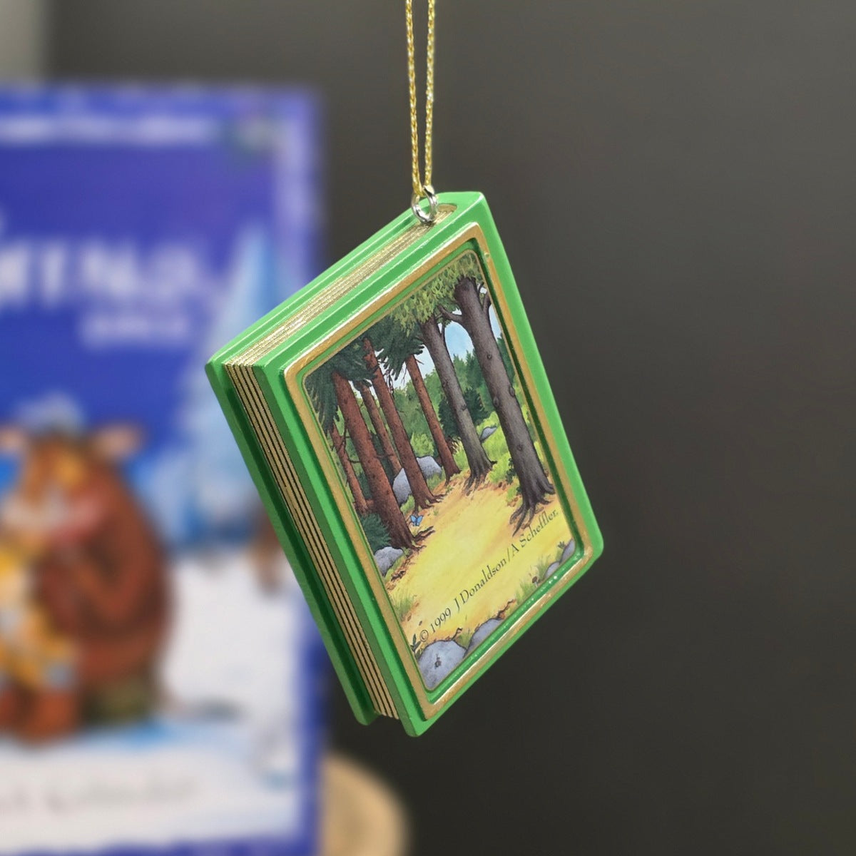 A green book-shaped ornament with a forest scene printed on the cover, hanging from a gold string against a dark blurred background.