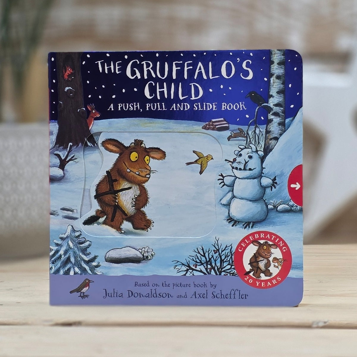 A children's push, pull and slide book titled 'The Gruffalo's Child' featuring an illustrated winter scene with The Gruffalo, a snowman, and a snowy landscape. The book is standing upright on a light wooden surface with a softly blurred indoor background.