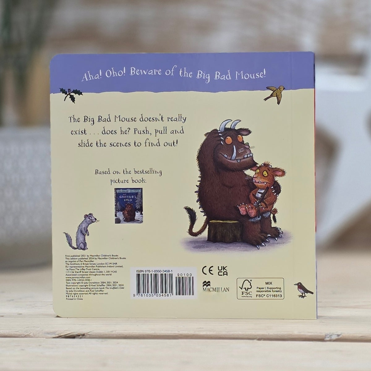 The back cover of a children's picture book titled 'The Big Bad Mouse' resting on a light wooden surface with a blurred outdoor background. The cover features a purple header, a beige background, and an illustration of a large brown mouse holding a small red mouse.