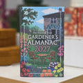 The National Trust Gardener's Almanac 2027 book standing upright on a light surface, with a colorful garden scene illustration on the cover featuring a house, flowers, chairs, and the title text.