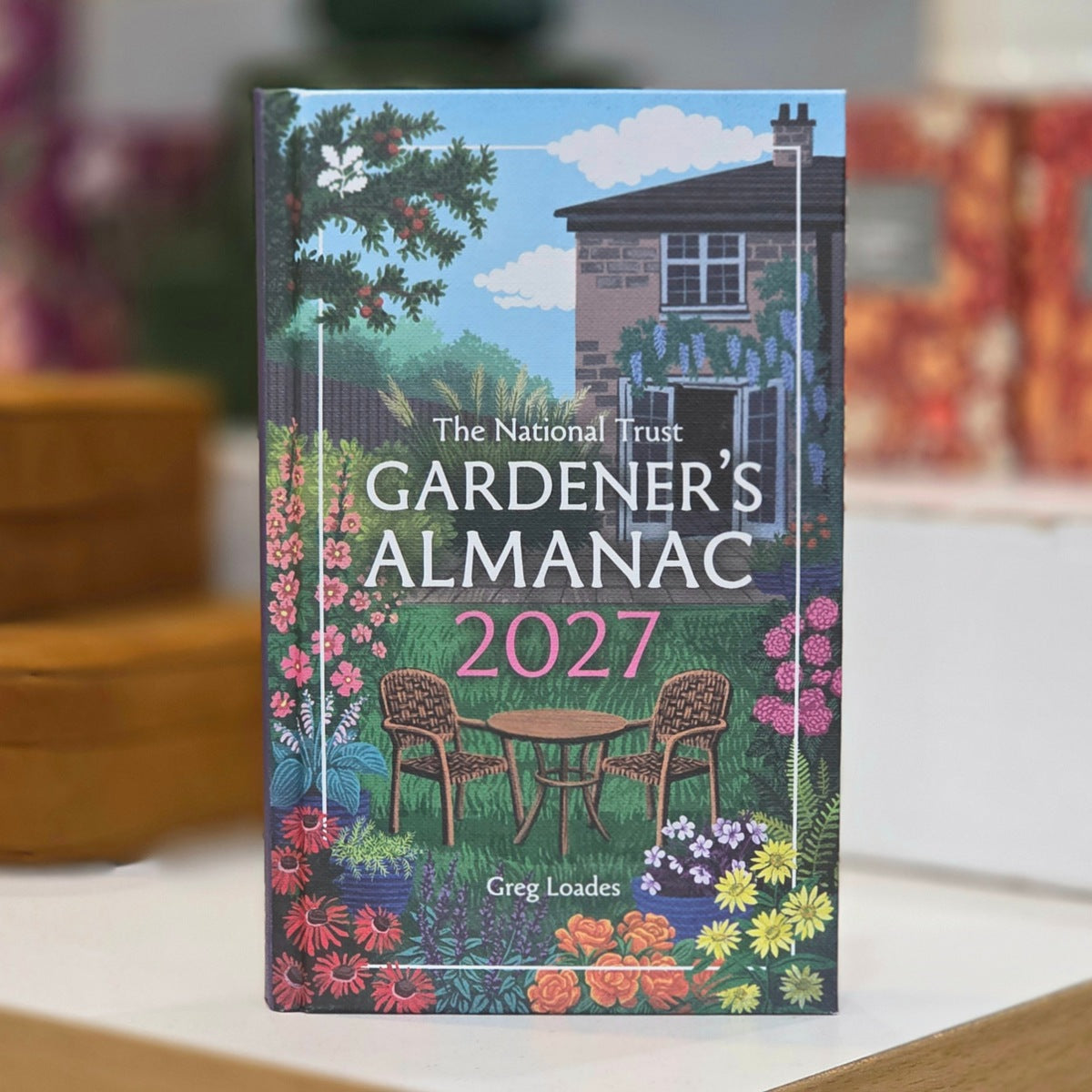 The National Trust Gardener's Almanac 2027 book standing upright on a light surface, with a colorful garden scene illustration on the cover featuring a house, flowers, chairs, and the title text.