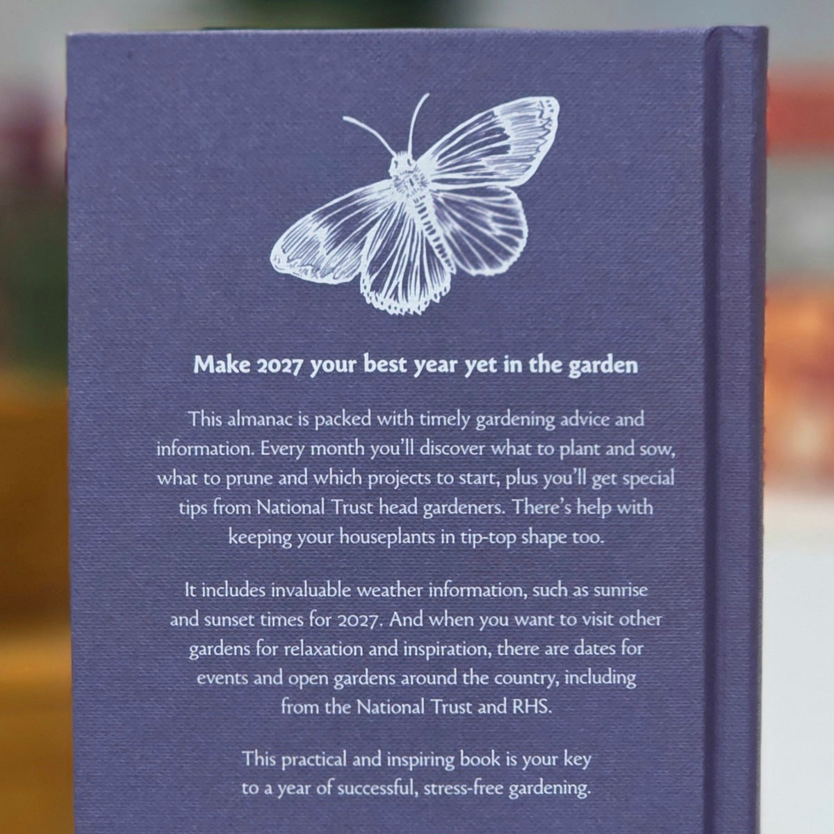 The back cover of a gardening book shown upright, featuring a white butterfly line drawing and descriptive text on a dark blue textured cover. The background is softly blurred with warm outdoor tones.