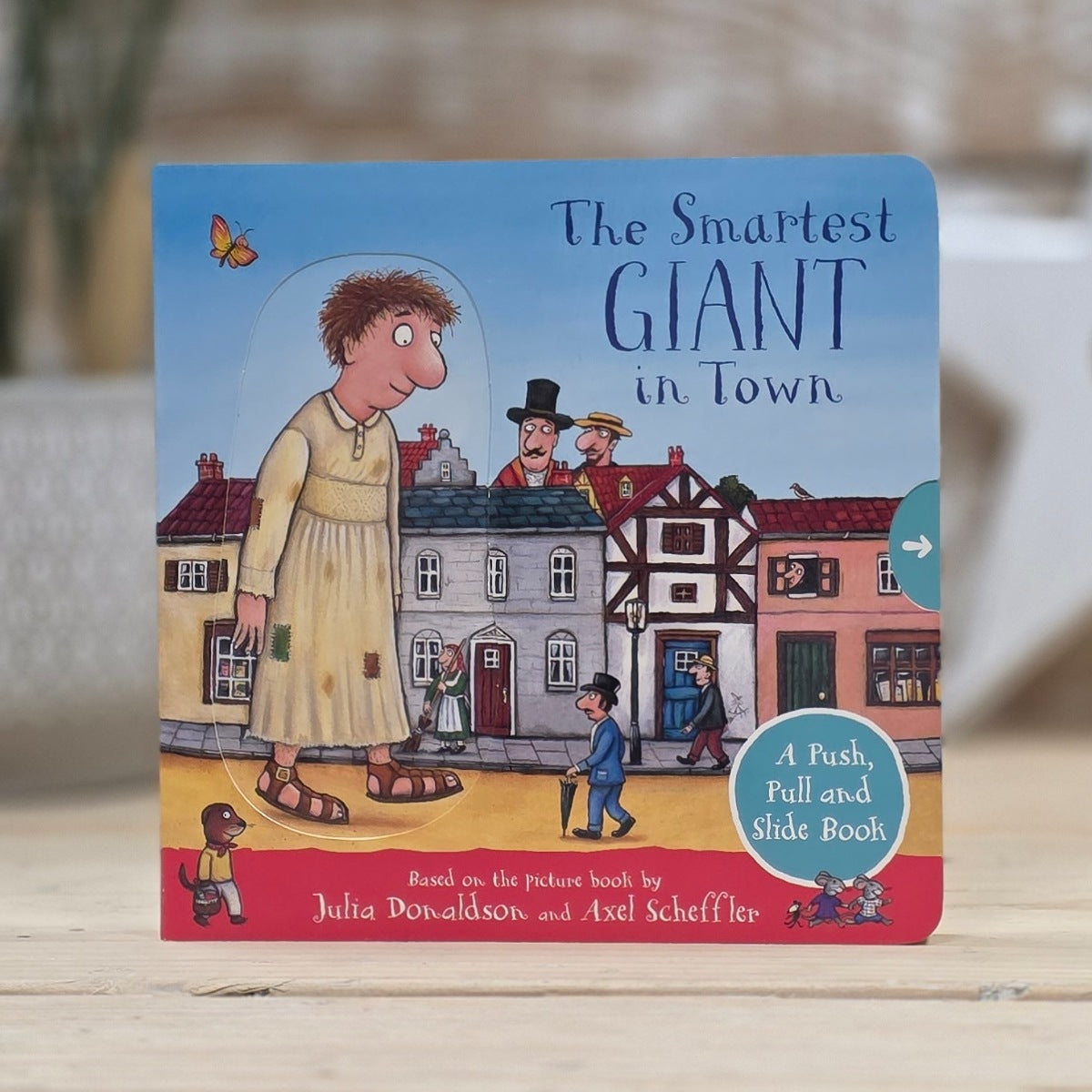 The Smarterest Giant in Town children's board book standing upright on a light wooden surface with a softly blurred indoor background. The cover shows an illustrated person in oversized clothing standing in front of old houses, with the title text 'The Smarterest Giant in Town' and a badge reading 'A Push, Pull and Slide Book.